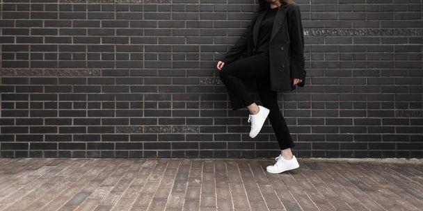 Cool girl in fashion denim pants in leather stylish sneakers in trendy jacket with black handbag standing on one leg near brick wall in city. Close-up female body. Fashionable casual youth womans wear - Photo, Image