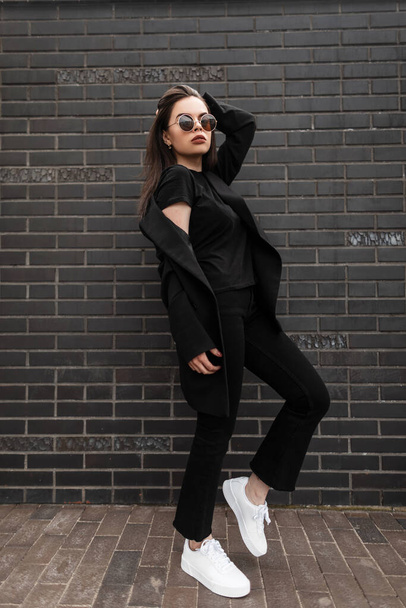 American modern νεαρή γυναίκα σε μοντέρνο μαύρο ντύσιμο σε κομψά γυαλιά ηλίου στη μόδα λευκά sneakers θέτει κοντά σε τούβλο κτίριο σε εξωτερικούς χώρους. Ελκυστικό κορίτσι σε νεανική casual στολή στην πόλη. Στυλ δρόμου - Φωτογραφία, εικόνα