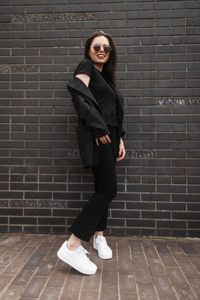 Modern young woman in fashionable black blazer in fashion t-shirt in stylish jeans pants stands near black brick wall in city. Close-up of female body. Summer collection of women's casual outfit. - Φωτογραφία, εικόνα