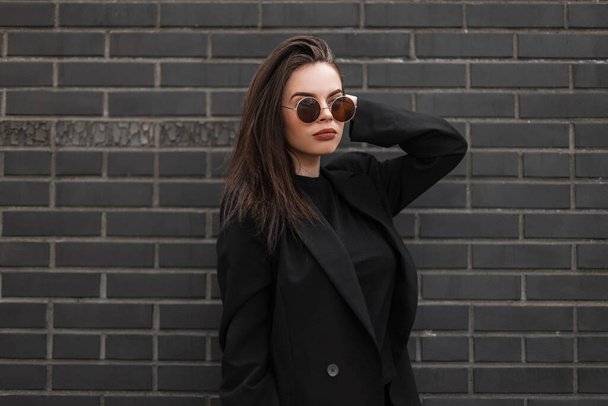 Fresh portrait young woman with sexy lips in round fashionable glasses in stylish black blazer in t-shirt near vintage building. Girl in trendy casual youth clothes posing near brick wall in street. - Foto, immagini