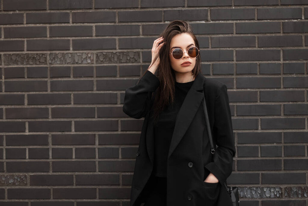 Divat modell gyönyörű fiatal nő hűvös kerek napszemüveg divatos fekete alkalmi kabát elegáns póló közelében modern téglafal a városban. Amerikai szexi lány divatos ifjúsági ruha az utcán. - Fotó, kép