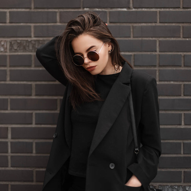 Stilvolle junge Frau mit jugendlicher Sonnenbrille in modischer schwarzer Kleidung mit Tasche posiert in der Nähe einer alten Backsteinmauer in der Stadt. Trendiges Modelmodel modernes Mädchen glättet die Haare und blickt nach unten. Straßenstil. - Foto, Bild