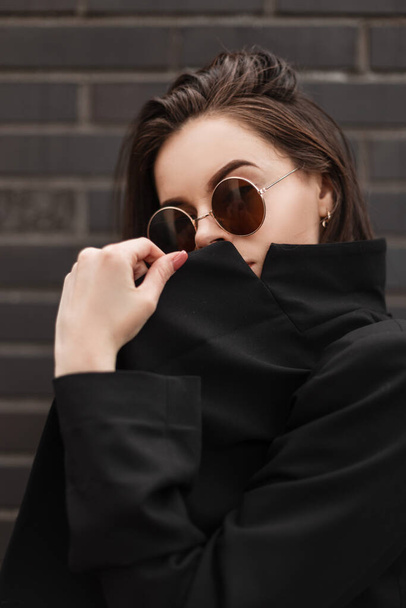 Fresh portrait young woman model in black casual youth clothes outdoors. American fashion girl in stylish round sunglasses in retro style closes face trendy jacket near black brick building in city. - Photo, Image