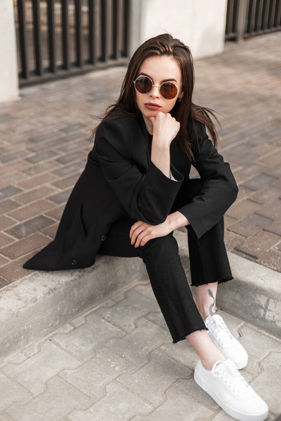 Fashion young woman in trendy pants in stylish youth blazer sitting on stone tiles in city. Fashionable new collection of women's clothing. Details of everyday look. Urban female business style - Photo, Image