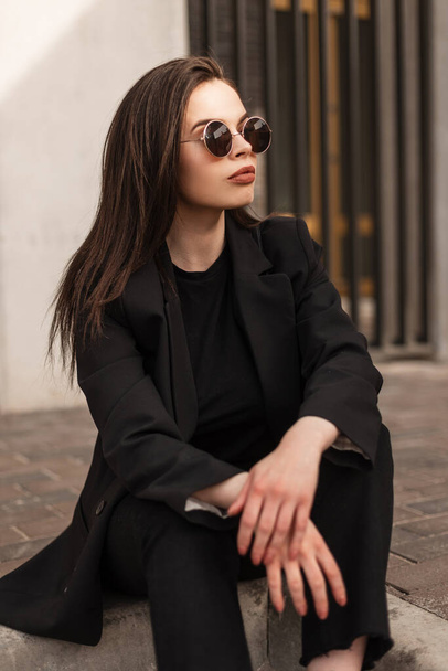 Şık tişörtlü, güneş gözlüklü, şık bir kadın model şehirde oturuyor. Siyah moda elbiseli manken kız sokaklarda seramik üzerinde dinleniyor. Kadınlar için şık kıyafetler.. - Fotoğraf, Görsel