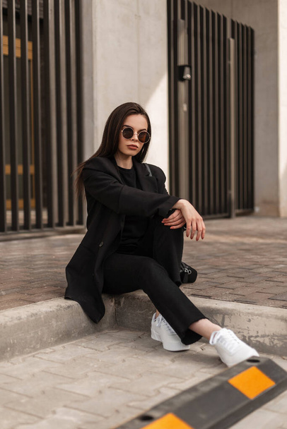 Wunderschöne junge Frau in modischer Sonnenbrille in stilvoller schwarzer Kleidung entspannen auf Steinfliesen in der Nähe moderner Gebäude in der Stadt. Amerikanisches Mädchen in lässigem Kleid ruht im Freien. Straßenstil. - Foto, Bild