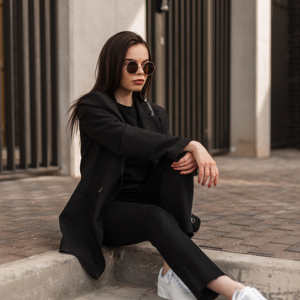 Прекрасна модна молода жінка в модних сонцезахисних окулярах в холодному чорному молодіжному одязі відпочиває на кам'яній плитці поблизу старовинної будівлі в місті. Модна дівчина модель моди в стильному вбранні сидить на вулиці
. - Фото, зображення