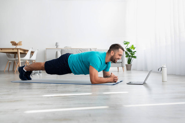 Happy young handsome guy athlete with beard do plank and watch fit lesson in living room - Fotoğraf, Görsel