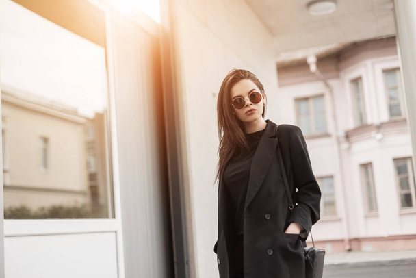 Preciosa mujer joven sexy en hermosa chaqueta negra en camiseta vintage en gafas de sol con estilo se encuentra cerca del edificio blanco al atardecer. Americana modelo de moda chica bastante de moda en el día soleado al aire libre. - Foto, imagen