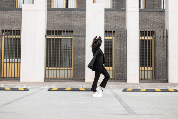 流行の革の白いスニーカーでスタイリッシュな黒の衣装でファッショナブルなかなり若い女性が街の近代的な建物の近くを歩く。都会の女の子のファッションモデルは通りを旅する。カジュアルユーススタイル。春の表情 - 写真・画像