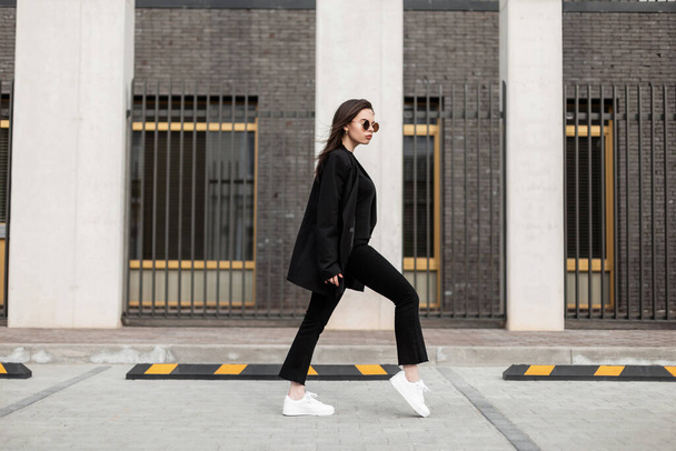 Cool νεαρή γυναίκα με γυαλιά ηλίου μόδας σε μοντέρνα μαύρα casual ρούχα σε λευκό μοντέρνα δερμάτινα sneakers περπατά κοντά στο σύγχρονο κτίριο. Ελκυστική ευρωπαϊκή κοπέλα σε casual στολή νεολαίας ταξιδεύει στην πόλη. - Φωτογραφία, εικόνα