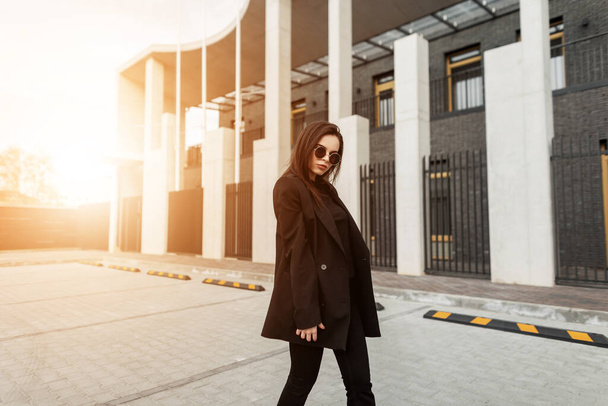 Schöne moderne Modell junge Frau in modischen lässigen schwarzen Kleid mit stilvoller Sonnenbrille reist Straße bei Sonnenuntergang. Schöne trendige Mädchen auf dem Hintergrund des orangen Sonnenlichts im Freien. Frühlings-Look. - Foto, Bild