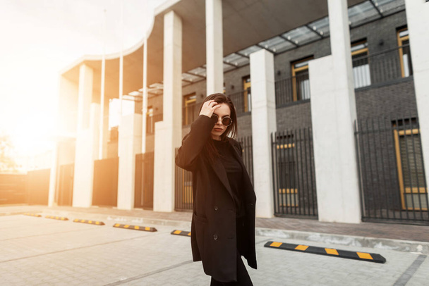 Fashionable young woman in stylish black blazer walks near building in city beautiful at sunset. Lovely gorgeous girl travels and enjoys sunbeams on street. Casual youth trendy clothes for women. - Фото, зображення