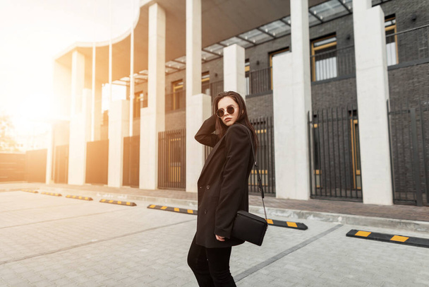 Fashionable young woman in stylish black blazer with vintage handbag walks near building in city at sunset. Urban girl travels and enjoys sunbeams on street. Casual youth trendy clothes for women. - Photo, Image