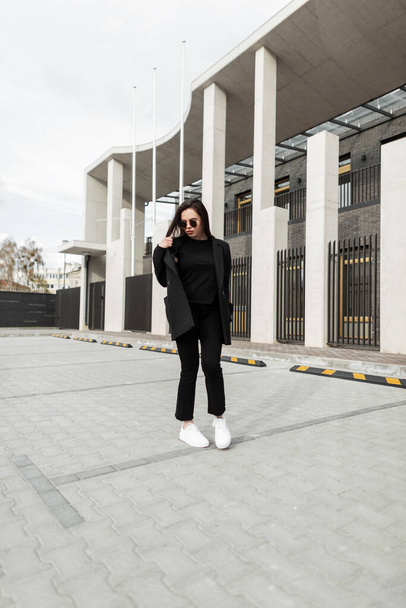 Muoti malli houkutteleva nuori nainen tyylikäs aurinkolasit muodikkaita mustia vaatteita uusi kesä trendikäs kokoelma on kävelee lähellä modernia rakennusta kaupungissa. Hieno tyttö vintage takki ulkona. - Valokuva, kuva