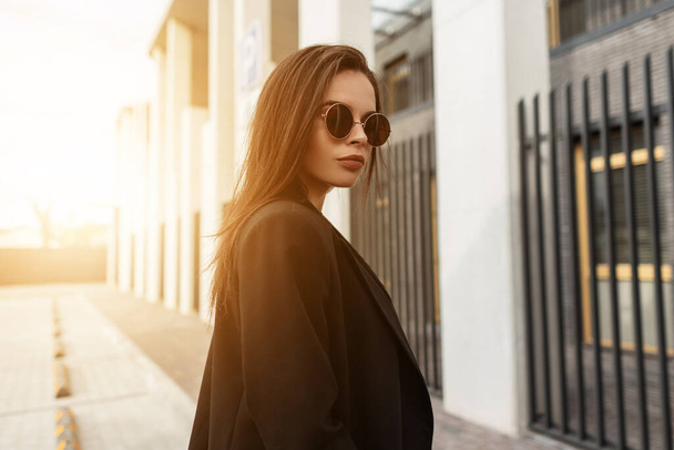 Sunny portrait beautiful young woman with sexy lips in youth fashionable sunglasses in stylish black clothes near modern building at sunset. Attractive girl model walks and enjoys orange sunlight. - Photo, Image