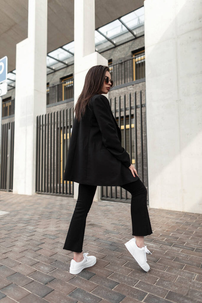 Модная молодая женщина в стильном винтажном черном пиджаке в модных джинсах стоит на улице. Современная девушка в молодежной повседневной куртке в городе. Леди в черном модном костюме на улице. Женский городской стиль - Фото, изображение