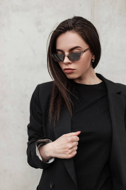 Стильный портрет сексуальная красивая молодая женщина в молодых модных солнцезащитных очках в черном винтажном пиджаке в модной футболке возле современной винтажной стены города. Привлекательная американская модель на улице - Фото, изображение