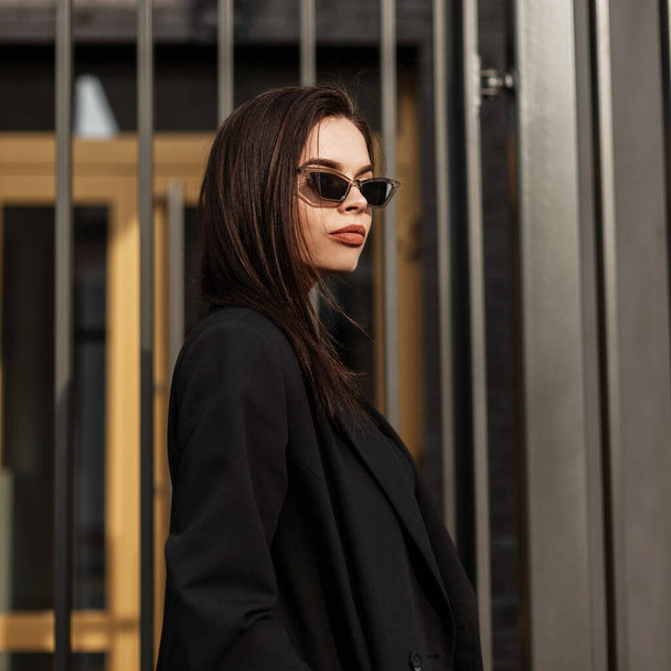 American stylish νεαρή γυναίκα με καστανά μαλλιά με σέξι χείλη σε μοντέρνα ρετρό γυαλιά ηλίου σε μαύρο vintage σακάκι στέκεται κοντά vintage μεταλλικό τοίχο στην πόλη. Σέξι μοντέρνα κοπέλα με μαύρα ρούχα στο δρόμο. - Φωτογραφία, εικόνα