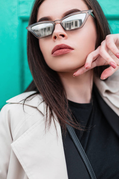 Modern fashion. Portrait woman in fashionable trench coat in black blazer in stylish T-shirt posing near turquoise wooden wall in street. New collection trendy clothes for women - 写真・画像