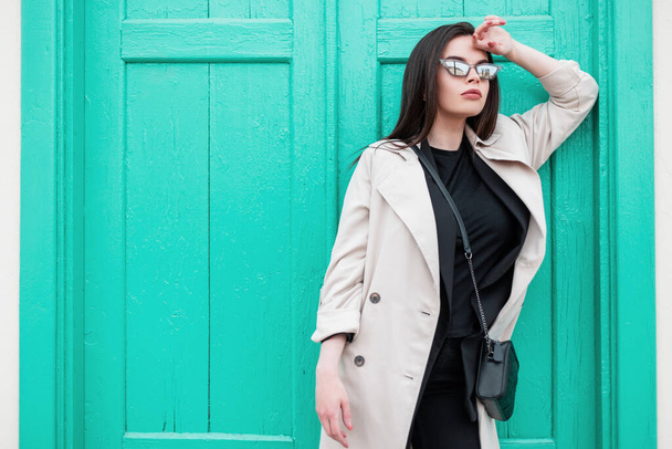 Sexy young woman in stylish retro sunglasses in black trendy t-shirt in fashionable spring trench coat is rest near vintage bright wooden turquoise door on street. Attractive girl model in the city. - Foto, imagen