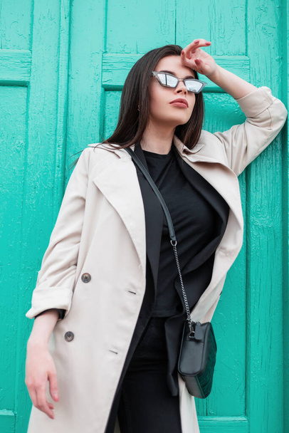Νεαρή γυναίκα σε ρετρό σακάκι σε μαύρο trendy t-shirt στη μόδα άνοιξη καμπαρντίνα υπόλοιπο κοντά vintage φωτεινό ξύλινο τυρκουάζ πόρτα στο δρόμο. Κορίτσι της μόδας σε casual ρούχα από κοντά. Στυλ νεολαίας. - Φωτογραφία, εικόνα