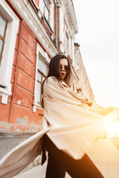 Coole junge Frau mit modischer Sonnenbrille im modischen frühlingshaften beigen Trenchcoat posiert bei Sonnenuntergang auf der Straße in der Nähe des Gebäudes. Ziemlich schöne urbane Mädchen Modell genießt Spaziergang und orange helles Sonnenlicht im Freien. - Foto, Bild