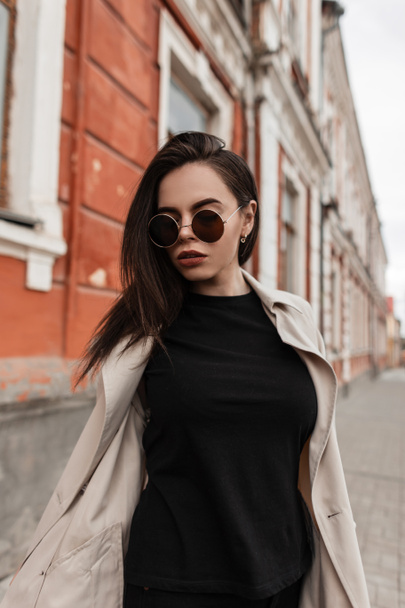 Stylish pretty urban young woman in black wear in fashionable spring youth trench coat in trendy sunglasses walk near vintage wall in street. Fine girl hipster posing outdoors in city. Casual look. - Photo, Image