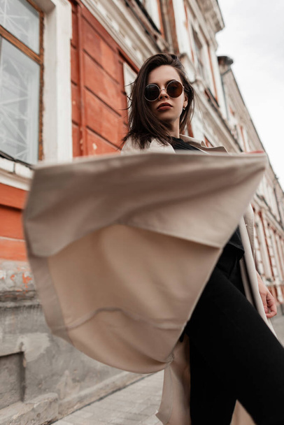 Americká mladá žena hipster v módní sluneční brýle v černé stylové ležérní džíny v jarní módní trenč kabát točí venku v blízkosti staré budovy na ulici. Městská dívka model v oblečení pro mládež ve městě. - Fotografie, Obrázek