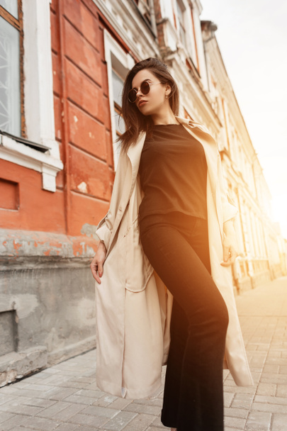 Elegante mujer joven urbana en ropa de moda en gafas de sol camina por la calle de la ciudad en el fondo naranja primavera brillante puesta de sol. Chica de moda en vestido casual disfruta de increíbles rayos sol y caminar al aire libre. - Foto, imagen