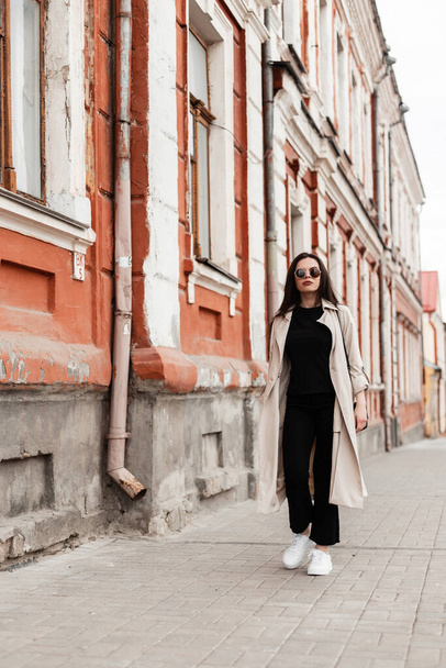 Stylish young woman in vintage sunglasses in fashionable spring clothes in white shoes with handbag walks in city near old building. Urban lovely girl hipster in trendy casual dress travels on street. - Foto, imagen