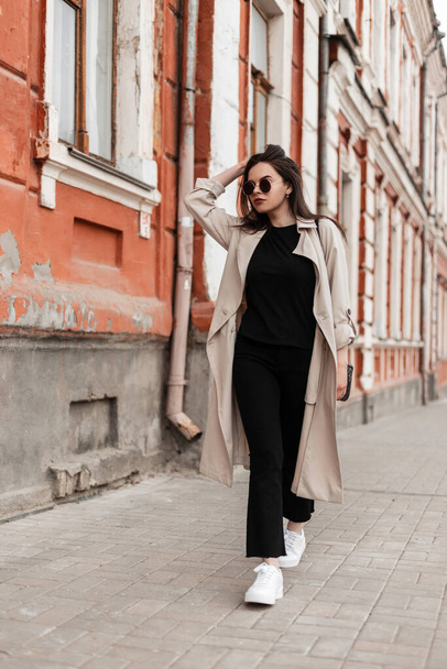 Urban gorgeous young woman in stylish sunglasses in casual spring clothes in white sneakers walks near vintage building in city. Lovely pretty trendy girl model walks outdoors. Street youth fashion. - Photo, Image