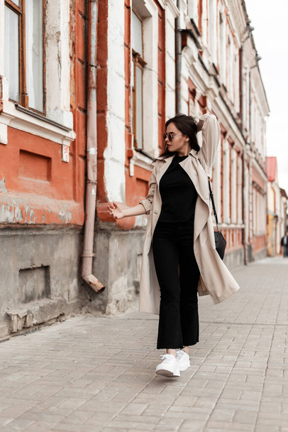 American young woman in stylish sunglasses in fashionable spring clothes in white shoes with handbag walks on street near vintage building. Lovely girl hipster in trendy casual dress travels in city. - Φωτογραφία, εικόνα