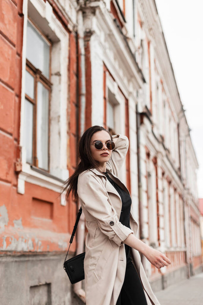 Modna młoda kobieta w zabytkowych okularach przeciwsłonecznych w stylowym płaszczu w czarnym ubraniu z modną skórzaną torbą w pobliżu starego budynku w mieście. Wspaniała modelka wychodzi na zewnątrz. Młodzież kobiet styl miejski. - Zdjęcie, obraz