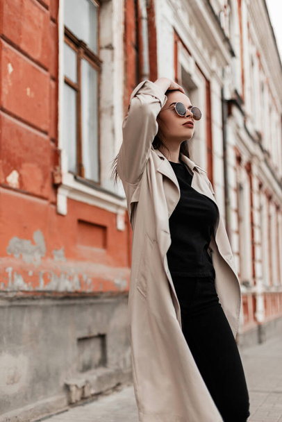 Hermosa joven hipster mujer joven en gafas de sol de moda en abrigo de trinchera vintage camina cerca del antiguo edificio de la ciudad. Precioso modelo de chica hermosa en ropa de abrigo casual de moda posa en la calle en el día de primavera. - Foto, Imagen