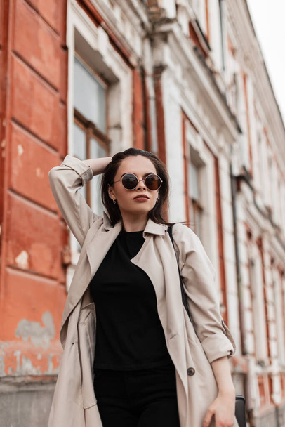 Pretty american young woman with brown hair in stylish trench coat posing standing outdoors in city. Urban beautiful stylish girl fashion model walks on street. Trendy seasonal women's outerwear. - Photo, Image