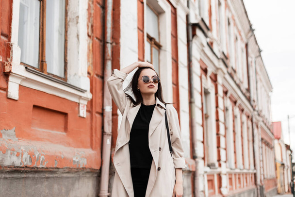 Gorgeous young woman in fashion black clothes in stylish sunglasses in beige trench coat walks and straightens hair. Attractive girl model in casual outfit walk on city street near vintage building. - Photo, Image