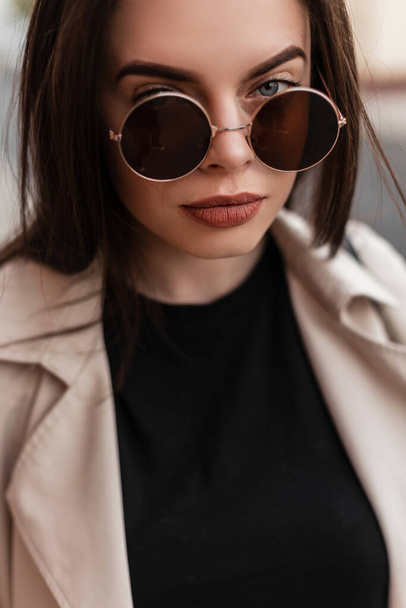 Fashion portrait beautiful young woman in round fashionable sunglasses in beige stylish trench with sexy lips outdoors in city. Gorgeous girl fashion model in casual vintage clothes. Street style. - Valokuva, kuva