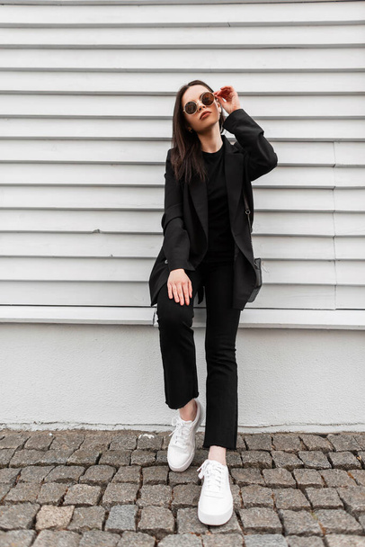 Csinos fiatal nő divat modell fehér vintage cipő elegáns fekete ruházat kézitáskával egyenesíti divat napszemüveg közelében fa épület a városban. Szép lány alkalmi ruhában pózol az utcán. - Fotó, kép