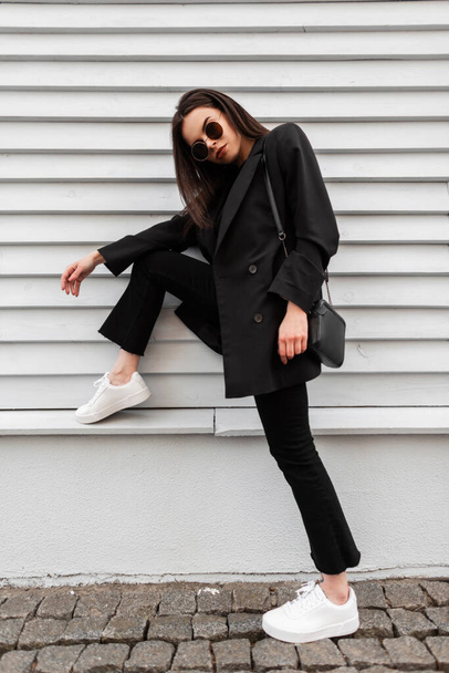 Modern fiatal nő divat modell divatos napszemüveg fehér cipőben elegáns fekete ruházat kézitáskával pihenni közelében faépület a városban. Vonzó lány alkalmi ruhában pózol a szabadban. - Fotó, kép