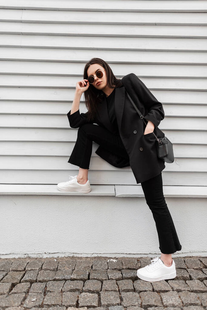 Pretty beautiful young woman in trendy sunglasses in cool black youth clothes in white trendy sneakers with stylish handbag is resting near vintage building in city. Fashionable girl enjoys walk. - Foto, immagini