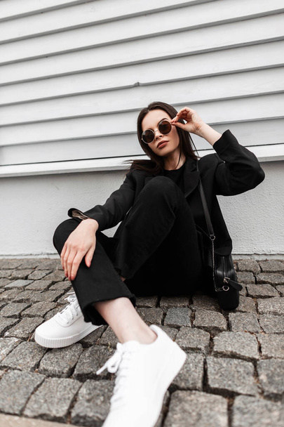 Gorgeous young woman in blazer in t-shirt in jeans in gym shoes with handbag in sunglasses sit near vintage wooden building on street. Beautiful girl in fashionable casual youth outfit poses outdoors. - 写真・画像