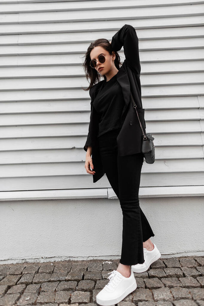 Stylish young woman in fashionable youth black clothes in sunglasses in white trendy sneakers with leather handbag poses near wooden building in city. American girl fashion model walks on street. - Fotó, kép