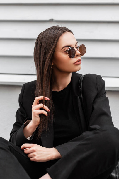 Trendy young woman hipster in stylish sunglasses in fashionable youth black clothes sit near wooden gray vintage wall outdoors. Attractive pretty gorgeous girl fashion model relaxes on street. - Foto, immagini