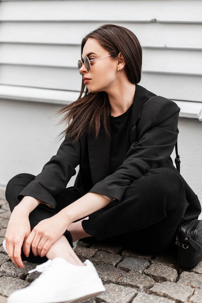 Городская молодая женщина модель в модных солнцезащитных очках в белых туфлях в стильной черной одежде с сумочкой отдохнуть возле деревянного здания в городе. Привлекательная девушка в повседневной одежде отдыхает на улице. - Фото, изображение