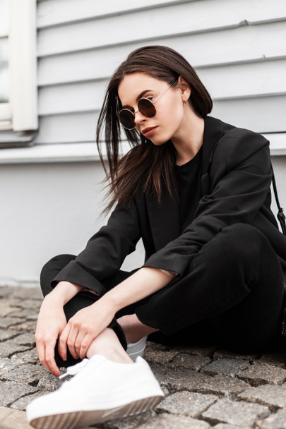 American νεαρή γυναίκα σε μοντέρνα γυαλιά ηλίου σε όμορφα μαύρα ρούχα της νεολαίας σε λευκό trendy sneakers υπόλοιπο σε πλακάκι πέτρα κοντά vintage κτίριο στην πόλη. Cool κορίτσι στη μόδα casual φθορά. Στυλ δρόμου. - Φωτογραφία, εικόνα