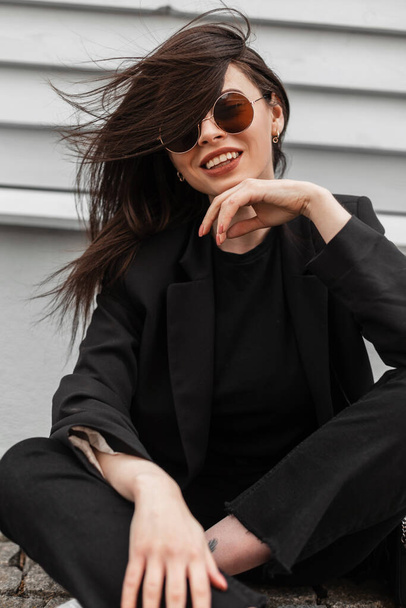 Улыбающаяся счастливая молодая женщина в модных солнцезащитных очках в красивой черной молодежной одежде отдыхает возле старинного здания в городе. Красивая позитивная девушка с красивой улыбкой в модной повседневной одежде сидит на улице - Фото, изображение