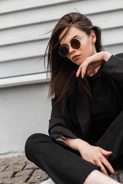 Trendy sexy urban young woman in fashionable round sunglasses in stylish black youth jacket enjoy a rest near vintage wooden wall in city. Beautiful modern girl fashion model in casual wear outdoors. - Foto, Imagen