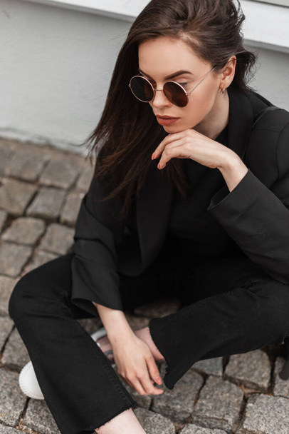 Young beautiful woman in trendy sunglasses in cool black youth clothes is resting near vintage building in city. Fashionable girl enjoys a rest on stone tile on street. Spring fashion wear for women. - Foto, Imagem