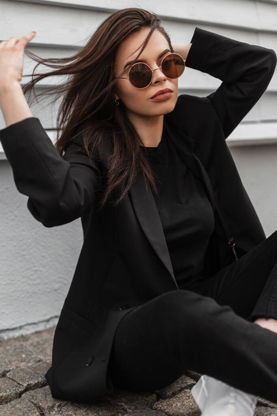 Pretty trendy young woman in fashion sunglasses in cool black youth clothes in white sneakers relaxes on stone tile near vintage building in city. Fashionable girl in stylish outfit poses in street. - Photo, Image